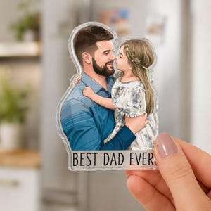 May include: A clear acrylic magnet featuring an illustration of a father and daughter embracing. The father wears a blue shirt, and the daughter wears a floral dress. The text "BEST DAD EVER" is at the bottom. The magnet is held by a hand.