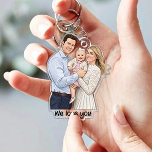 May include: A clear acrylic keychain featuring a family portrait of a man, woman, and baby. The man wears a blue shirt, the woman a white dress, and the baby a pink outfit. The keychain has a silver ring and the text "We love you".