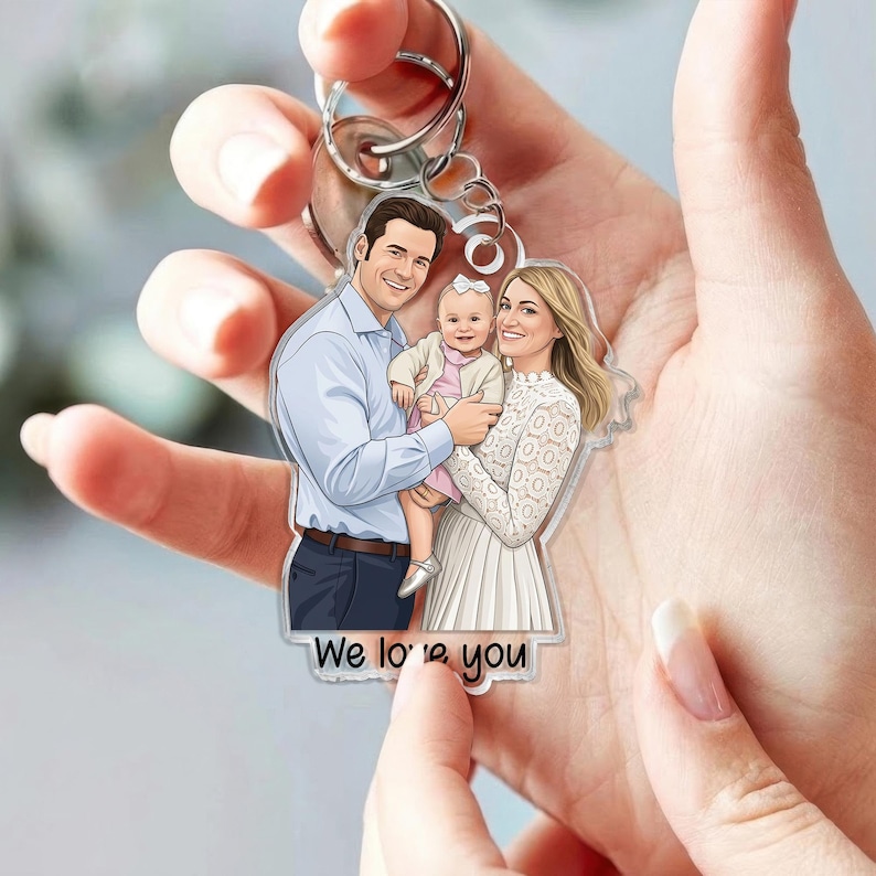 May include: A clear acrylic keychain featuring a family portrait of a man, woman, and baby. The man wears a blue shirt, the woman a white dress, and the baby a pink outfit. The keychain has a silver ring and the text "We love you".
