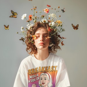 Puede incluir: Una persona con una camiseta blanca con el texto "BREAKFAST WITH A SIDE OF NOQUISITIC" y un dibujo de una mujer sosteniendo panqueques. Mariposas y flores adornan su cabeza, con mariposas en el fondo.