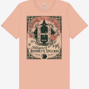 K&ouml;nnte beinhalten: Ein pfirsichfarbenes T-Shirt mit einer detaillierten Grafik. Das Design zeigt einen gro&szlig;en Buchstaben "H" mit floralen Akzenten, den Namen "Hilda" und den Text "Hildegard Antoinette Spellman". Am unteren Rand befindet sich ein kleiner Kessel.