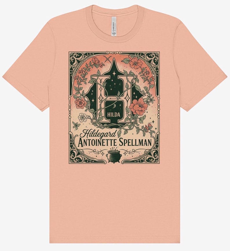 K&ouml;nnte beinhalten: Ein pfirsichfarbenes T-Shirt mit einer detaillierten Grafik. Das Design zeigt einen gro&szlig;en Buchstaben "H" mit floralen Akzenten, den Namen "Hilda" und den Text "Hildegard Antoinette Spellman". Am unteren Rand befindet sich ein kleiner Kessel.