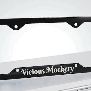 May include: A rectangular black license plate frame with rounded corners and four mounting holes. The bottom center of the frame features the white text Vicious Mockery in a stylized serif font, centered between two small white circular accents.