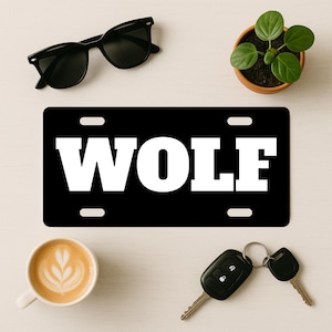 May include: Black license plate with the word "WOLF" in bold white letters. The plate has four mounting holes. Sunglasses, a small potted plant, a cup of coffee, and car keys are also visible.
