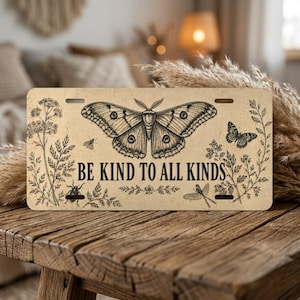 May include: A license plate with a vintage design featuring a large moth, a butterfly, and the text "BE KIND TO ALL KINDS." The plate has a beige background with botanical illustrations and insect drawings.