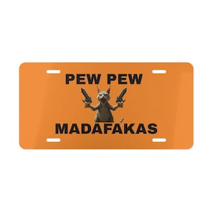 May include: Orange license plate with the words "PEW PEW MADAFAKAS" in black. A cat holding two guns is in the center. The license plate has four mounting holes, one in each corner.