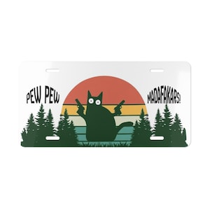 May include: A white license plate featuring a black cat silhouette holding two guns, set against a sunset backdrop. The plate includes the phrases "PEW PEW" and "MADAF*KARS!" with green trees on the bottom.