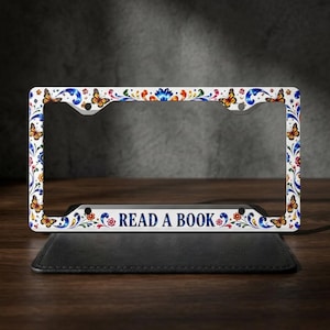 May include: A rectangular license plate frame featuring a white background with a colorful pattern of orange butterflies, red flowers, and blue swirling floral motifs. The bottom center of the frame displays the text READ A BOOK in blue.