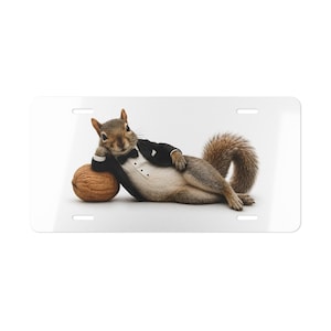 May include: A rectangular white license plate featuring a graphic of a brown squirrel wearing a black tuxedo jacket with a white shirt and a black bow tie. The squirrel is lying on its side, resting its head on a large brown textured walnut.