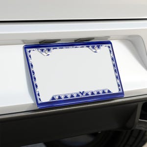 May include: A blue and white license plate frame with a decorative border. The frame is rectangular and has a white center with a blue patterned border. The frame is mounted on a white vehicle.