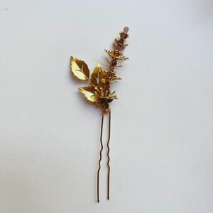 K&ouml;nnte beinhalten: Goldfarbene Haarnadel mit floralem Design. Die Nadel hat stilisierte goldene Bl&auml;tter und einen Stiel, der mit kleinen lila Perlen verziert ist. Der Text "Style 1" ist unten aufgedruckt.