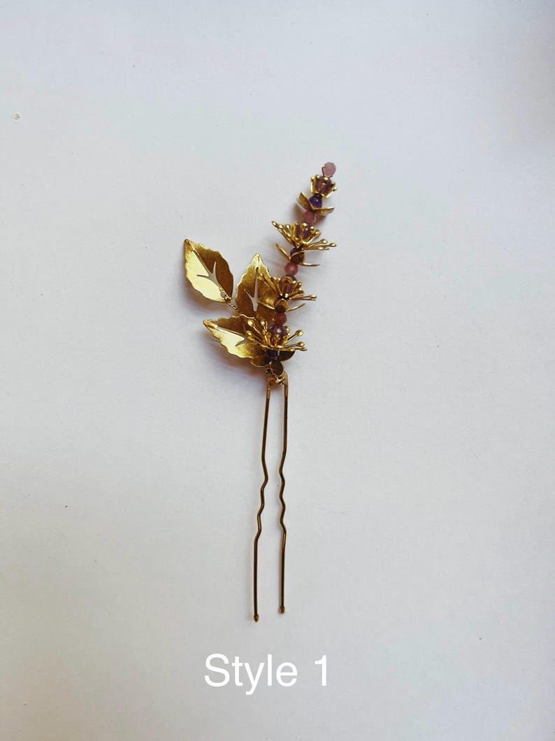 K&ouml;nnte beinhalten: Goldfarbene Haarnadel mit floralem Design. Die Nadel hat stilisierte goldene Bl&auml;tter und einen Stiel, der mit kleinen lila Perlen verziert ist. Der Text "Style 1" ist unten aufgedruckt.
