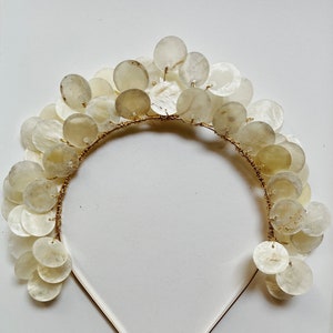 Könnte beinhalten: Ein goldenes Stirnband mit einer Reihe von weißen, runden, muschelförmigen Ornamenten. Die Ornamente sind in einer kaskadierenden Anordnung angeordnet.