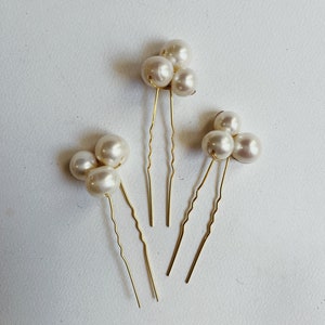 May include: Three gold hair pins with pearl clusters. Each pin has three pearls, and the pearls are white and round.