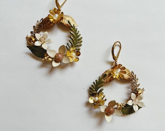 Può includere: Un paio di orecchini dorati con un design floreale. Gli orecchini sono in metallo e presentano fiori bianchi e gialli, foglie verdi e una perla marrone.