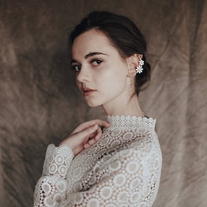 Może przedstawiać: Kobieta o ciemnych włosach w białej koronkowej bluzie z wysokim dekoltem. Ma na sobie delikatny kwiatowy kolczyk.