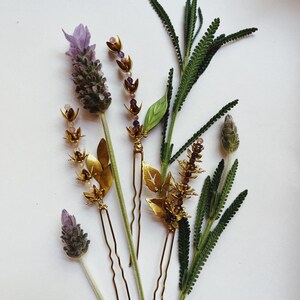K&ouml;nnte beinhalten: Draufsicht auf Haarschmuck und Lavendelzweige. Die Haarnadeln sind goldfarben mit Blatt- und Blumenmotiven. Der Lavendel hat violette Knospen und gr&uuml;ne Stiele. Der Hintergrund ist wei&szlig;.