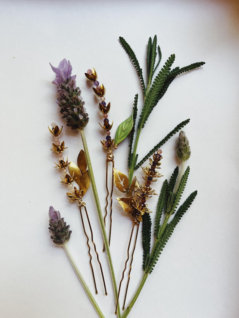 K&ouml;nnte beinhalten: Draufsicht auf Haarschmuck und Lavendelzweige. Die Haarnadeln sind goldfarben mit Blatt- und Blumenmotiven. Der Lavendel hat violette Knospen und gr&uuml;ne Stiele. Der Hintergrund ist wei&szlig;.