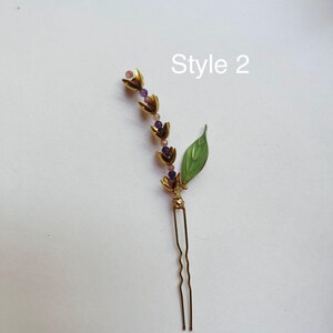 K&ouml;nnte beinhalten: Eine goldfarbene Haarnadel mit floralem Design, mit lila Perlen und einem gr&uuml;nen Blattakzent. Der Text "Style 2" ist sichtbar. Dieses dekorative Haaraccessoire eignet sich f&uuml;r verschiedene Frisuren.