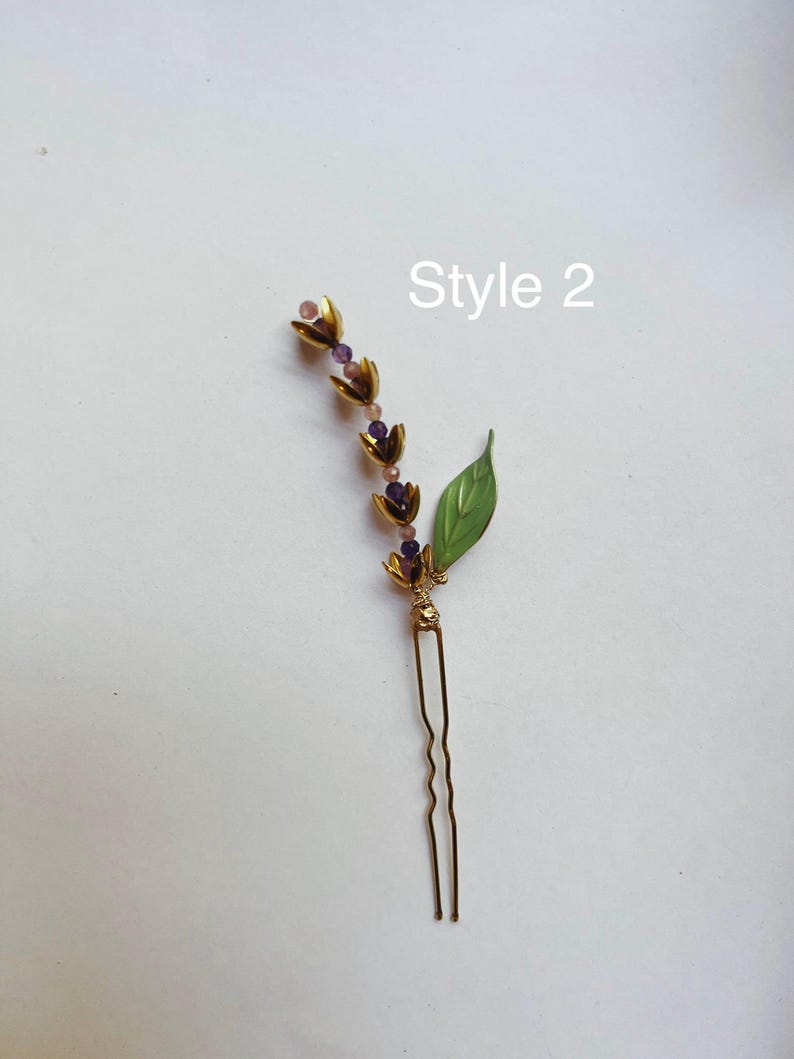 K&ouml;nnte beinhalten: Eine goldfarbene Haarnadel mit floralem Design, mit lila Perlen und einem gr&uuml;nen Blattakzent. Der Text "Style 2" ist sichtbar. Dieses dekorative Haaraccessoire eignet sich f&uuml;r verschiedene Frisuren.