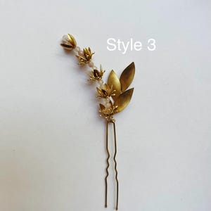 K&ouml;nnte beinhalten: Eine goldfarbene Haarnadel mit floralen und Blatt-Akzenten. Die Nadel hat kleine, detaillierte Bl&uuml;ten und Bl&auml;tter mit einer Ansammlung von helllavendelfarbenen Perlen. Der Text "Style 3" und "(Helllavendelfarben)" sind ebenfalls sichtbar.