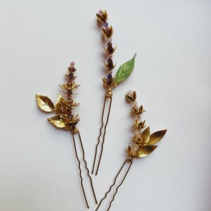 K&ouml;nnte beinhalten: Drei goldfarbene Haarnadeln mit Blumen- und Blattmotiven. Die Nadeln haben kleine lila und rosa Perlen und ein gr&uuml;nes Blatt. Die Haaraccessoires sind auf einer wei&szlig;en Oberfl&auml;che angeordnet.