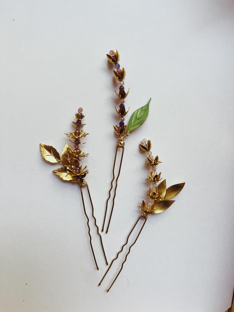 K&ouml;nnte beinhalten: Drei goldfarbene Haarnadeln mit Blumen- und Blattmotiven. Die Nadeln haben kleine lila und rosa Perlen und ein gr&uuml;nes Blatt. Die Haaraccessoires sind auf einer wei&szlig;en Oberfl&auml;che angeordnet.
