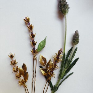 K&ouml;nnte beinhalten: Draufsicht auf drei goldfarbene Haarnadeln mit Blumen- und Blattmotiven, verziert mit kleinen lila und rosa Perlen. Ein Zweig Lavendel mit lila Bl&uuml;ten und gr&uuml;nen Bl&auml;ttern liegt neben dem Haarschmuck.
