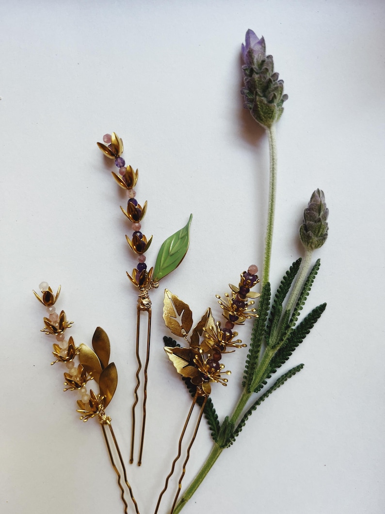 K&ouml;nnte beinhalten: Draufsicht auf drei goldfarbene Haarnadeln mit Blumen- und Blattmotiven, verziert mit kleinen lila und rosa Perlen. Ein Zweig Lavendel mit lila Bl&uuml;ten und gr&uuml;nen Bl&auml;ttern liegt neben dem Haarschmuck.