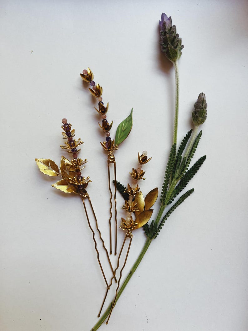 K&ouml;nnte beinhalten: Goldfarbene Haarnadeln mit Blumen- und Blattmotiven, verziert mit kleinen lila und rosa Perlen. Die Haarnadeln sind zusammen mit Lavendelzweigen angeordnet und zeigen ein nat&uuml;rliches, botanisches Thema. Ideal f&uuml;r Hochzeiten oder besondere Anl&auml;sse.