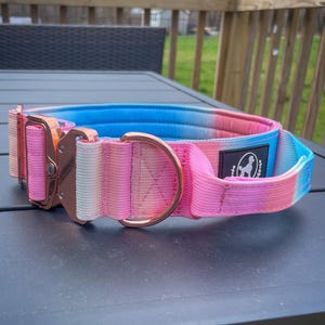 May include: A vibrant dog collar with a tie-dye design in pink, blue, and white. It has a rose gold metal buckle and a matching D-ring. A small black tag with a white logo is attached to the collar.
