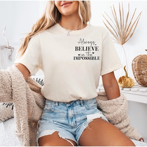 Könnte beinhalten: Cremefarbenes T-Shirt mit dem Text "Always Believe in the Impossible" in schwarzer Schrift. Das Shirt wird mit Jeansshorts kombiniert. Ein beigefarbener Cardigan ist über die Schultern gelegt. Ein dekorativer Kürbis und ein Weidenkorb sind im Hintergrund.