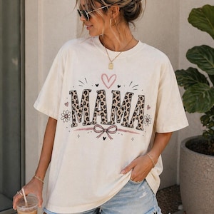 Könnte beinhalten: Cremefarbenes T-Shirt mit dem Wort "MAMA" im Leopardenmuster, einem rosa Herz und einer Schleife. Das Shirt wird mit Jeansshorts kombiniert. Das Bild zeigt einen lässigen, alltäglichen Stil.