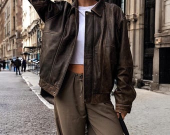 May include: A brown leather jacket with a zipper closure and a collared neckline. The jacket has a relaxed fit and is worn over a white crop top and brown pants.