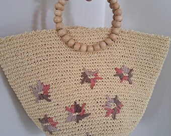 Puede incluir: Un bolso tote tejido beige con un asa de cuentas de madera. La bolsa está decorada con detalles florales en tonos rosa y gris. La bolsa está sujeta por una mano.