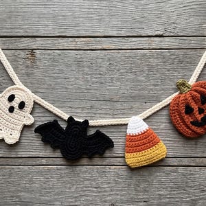 May include: A Halloween-themed crochet garland featuring four hanging ornaments attached to a cream-coloured cord. The ornaments include a white ghost with black eyes, a black bat, a piece of candy corn, and an orange pumpkin with a green stem.