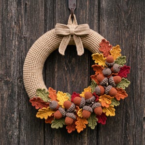 May include: A circular wreath made of tan crocheted yarn featuring a matching bow at the top. The lower right section is decorated with an arrangement of crocheted oak leaves in shades of red, orange, yellow, and green, interspersed with brown acorns.