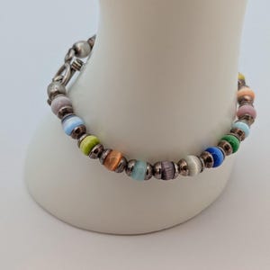 May include: A bracelet featuring a series of multicolored cat's eye beads separated by small, dark metallic spacers. The beads display a variety of hues including blue, green, orange, purple, and pink, and the bracelet secures with a metal clasp.