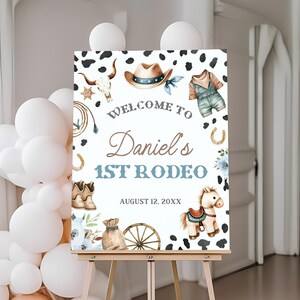 May include: A sign welcoming guests to Daniel's 1st Rodeo, decorated with cowboy-themed illustrations. The sign includes a cowboy hat, horseshoe, boots, and a toy horse. The sign is on a wooden easel, with cow print accents.