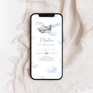 以下が含まれることがあります： 「ONE」と書かれた青い飛行機が白い雲でいっぱいの空を飛んでいます。飛行機の下には「マーティンは1歳になります。一緒に祝いましょう」という文字が印刷されています。招待状には、パーティーの日時と場所が記載されています。