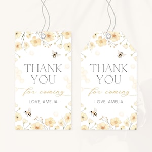 May include: Two white gift tags with floral and bee illustrations. The tags read "THANK YOU for coming, LOVE, AMELIA" in a mix of fonts. The tags are decorated with yellow flowers and bees, and have a string attached.