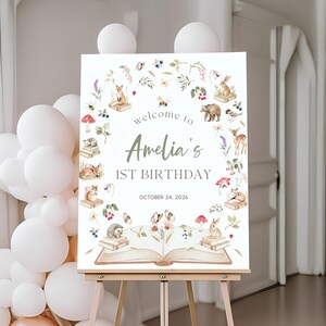 以下が含まれることがあります： アメリアの1歳の誕生日のウェルカムサイン。水彩画の森の動物と花のイラストが特徴です。「Welcome to Amelia's 1st Birthday」というテキストと2026年10月24日の日付が含まれています。木製のイーゼルに表示されています。