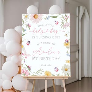 Op de afbeelding: Een welkomstbord voor Amelia's 1e verjaardag, weergegeven op een houten ezel. Het bord toont bloemen- en lieveheersbeestjes illustraties met de tekst "Our Little Ladybug is Turning One!" en de datum 25.08.2026.