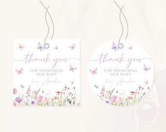 Op de afbeelding: Twee cadeaulabels voor een babyshower, één vierkant en één rond, met een bloemen- en vlinderontwerp. De labels bevatten de tekst "bedankt voor het douchen van onze baby" en een naam. De labels zijn wit met een touwtje eraan.