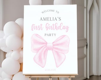 May include: A white sign on an easel with the text "WELCOME TO AMELIA'S first Birthday PARTY" in black and pink script. A large pink bow graphic is below the text. The date "12.08.2025" is at the bottom. White balloons are in the background.