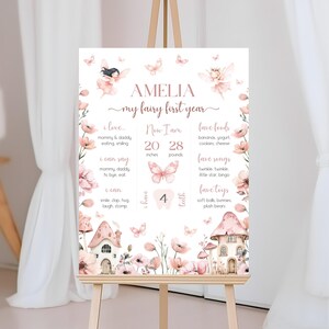 May include: A pink and white printable first birthday milestone chart with a fairy theme. The chart includes the child's name, Amelia, and details about their first year, including their height, weight, favourite foods, songs, and toys. The chart is decorated with butterflies, flowers, and a whimsical fairy house.