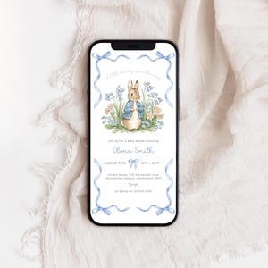 May include: A mobile phone shows a baby shower invitation with a watercolour illustration of a bunny in a blue jacket amidst flowers. The invitation includes the name "Olivia Smith" and event details, framed by blue ribbons.