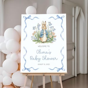 May include: A baby shower welcome sign featuring a watercolor illustration of a rabbit in a blue jacket surrounded by flowers. The sign reads "Welcome to Olivia's Baby Shower" with the date August 12, 2025, framed by blue ribbons and bows.