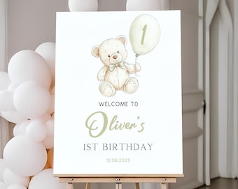 May include: A white sign on an easel with a watercolor illustration of a teddy bear holding a balloon with the number 1. The sign reads "Welcome to Oliver's 1st Birthday" and the date 12.08.2025.