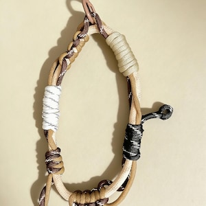 Könnte beinhalten: Eine mehrfarbige Hundeleine aus Seil mit beigen, braunen, schwarzen und weißen Abschnitten. Die Leine hat einen Metallverschluss und eine Schlaufe am Ende. Das Seil ist in verschiedenen Mustern geknotet und geflochten.